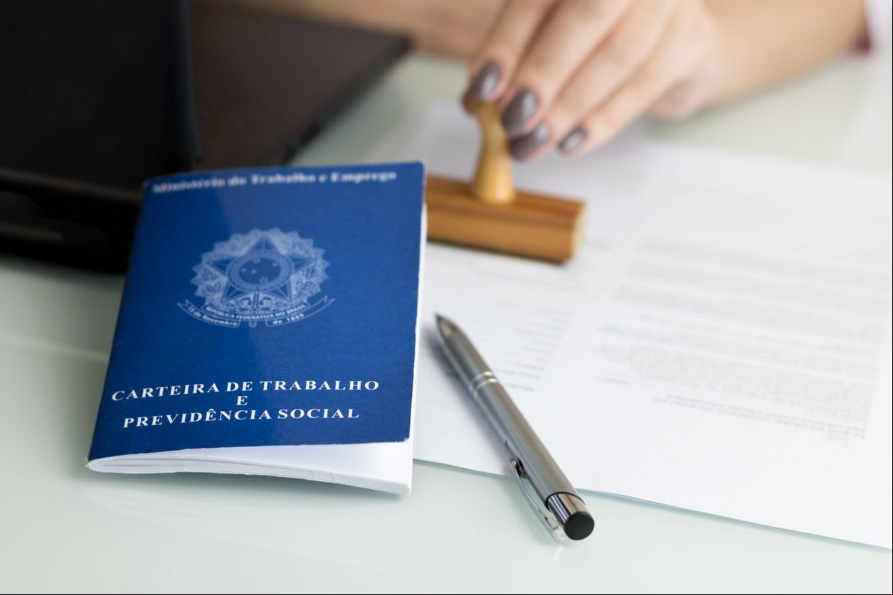 A portfolio of Brazilian workers (Translation "Work Permit, Ministry of Labor and Social Security Brazil CTPS"), female hand stamping a work contract background.