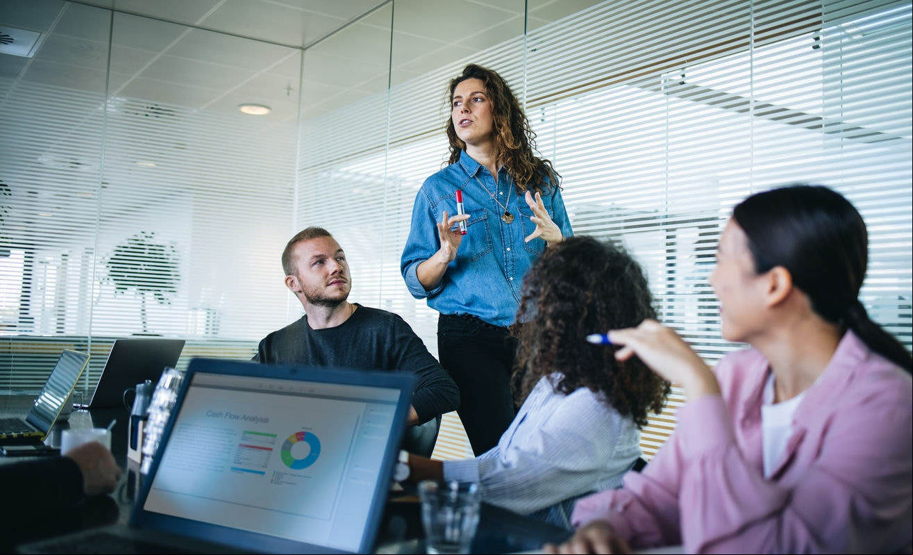 Businesswoman walking in office boardroom and sharing a strategy with team sitting at table. Female manager explaining new business plan with coworkers sitting at conference table in office.